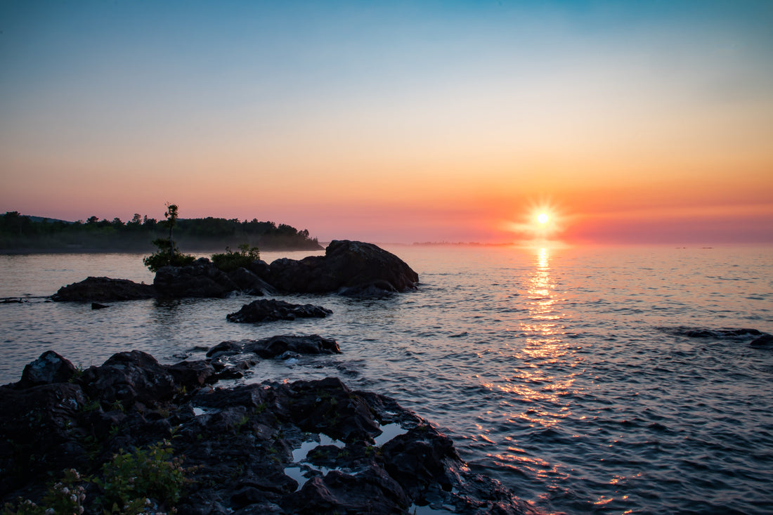 Sunrise over calm water with rocky shoreline, soft light breaking through early morning mist.