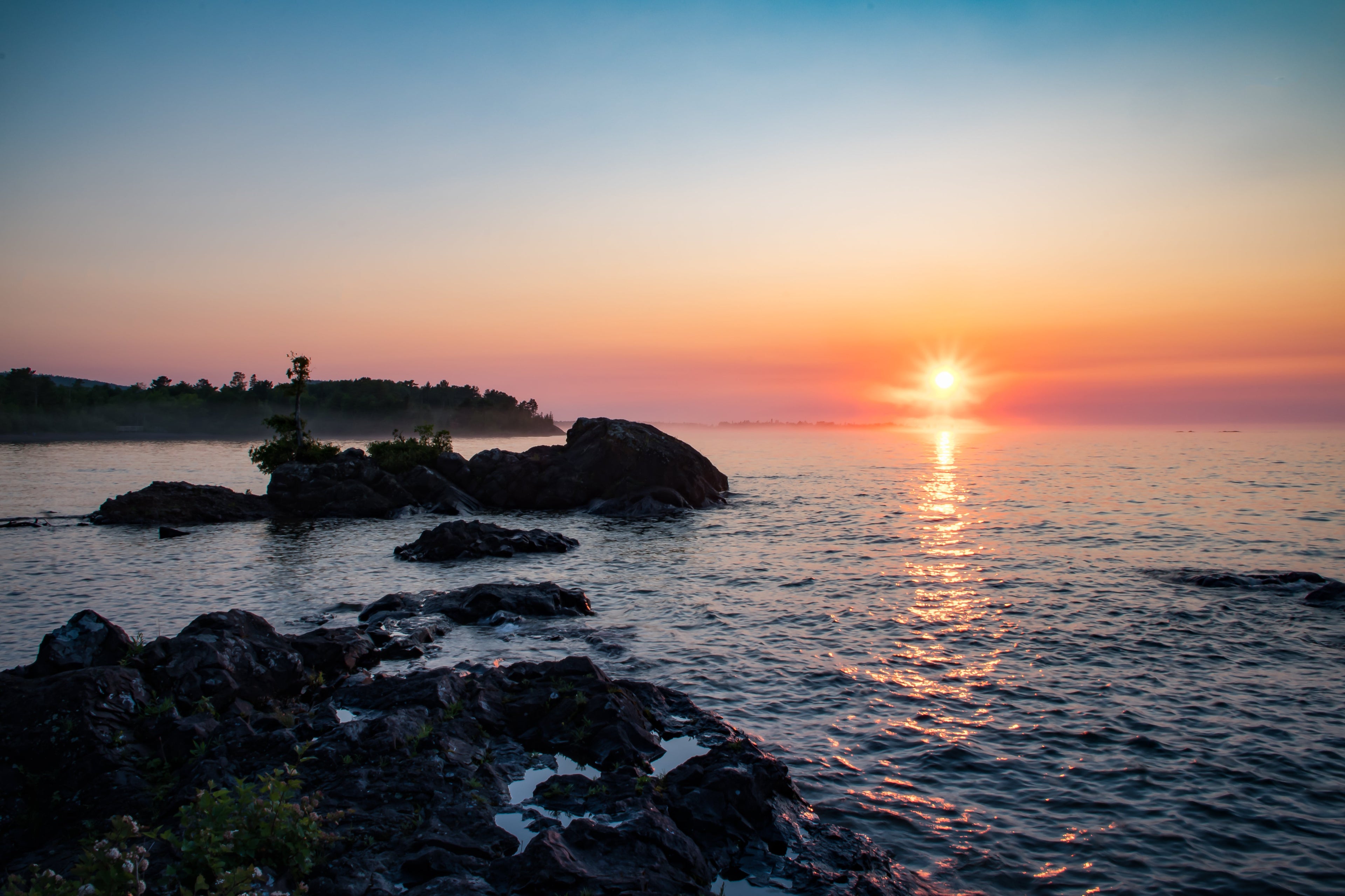 Sunrise over calm water with rocky shoreline, soft light breaking through early morning mist.